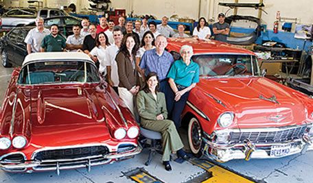 The Bertolucci's team gathered in the shop around a red 1962 Chevrolet Corvette and a red and white 1956 Chevrolet Bel Air convertible