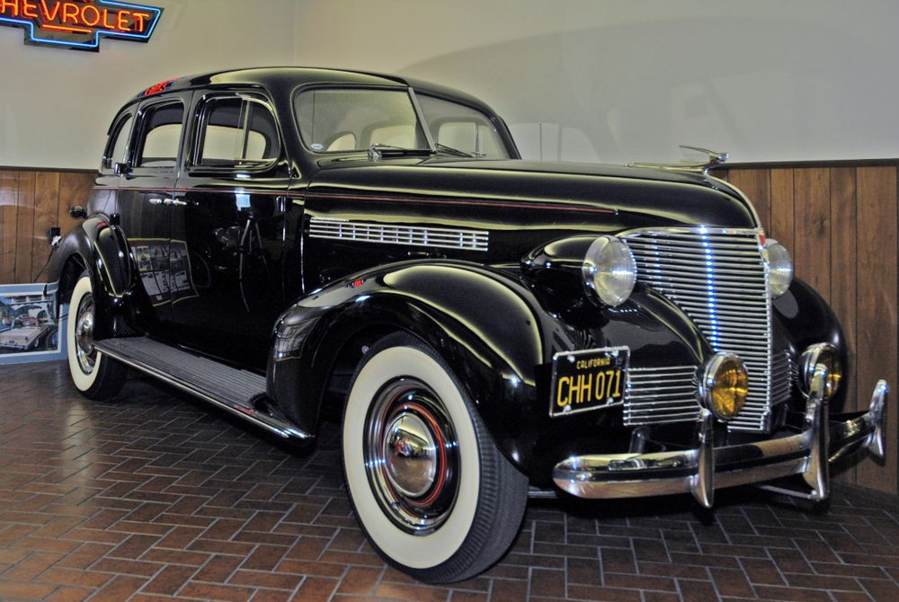 Black 1939 Chevrolet 4-door sedan with whitewall tires and yellow fog lights under a neon Chevrolet sign