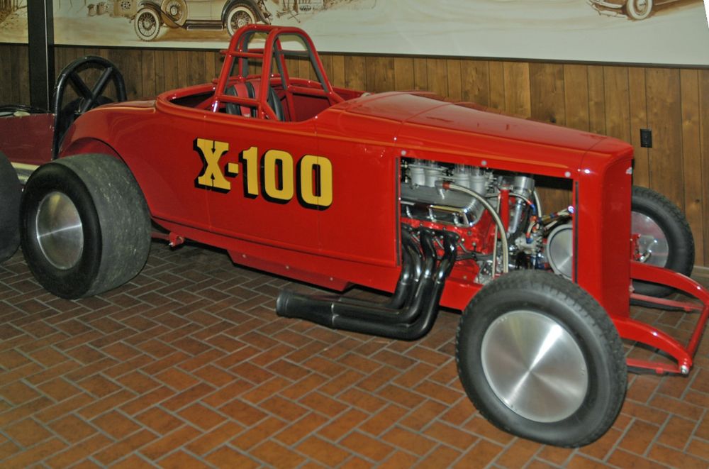 Red X-100 1930 Chevrolet roadster drag car with yellow X-100 lettering, roll bar, exposed V8 engine and black headers